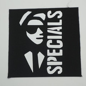 May include: A square black fabric patch featuring a white screen-printed graphic of a person wearing a hat and sunglasses, with the word SPECIALS printed vertically in bold white capital letters along the right side of the design.