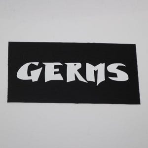 May include: A black rectangular patch with the word "GERMS" in bold, white, stylized lettering. The patch has a textured surface and is set against a white background.