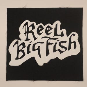 May include: A black square patch with the words "Reel Big Fish" in white, stylized lettering. The text is in a curved, flowing design, set against a solid black background. The patch has a slightly textured appearance.