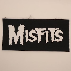 May include: A black rectangular patch with the word "MISFITS" in large, white, stylized lettering. The patch has a textured surface and is likely made of fabric.