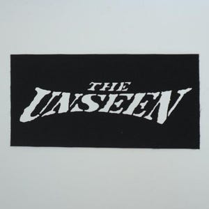 May include: A black rectangular patch with the white text "THE UNSEEN" in a stylized font. The patch has a textured surface and is set against a white background.