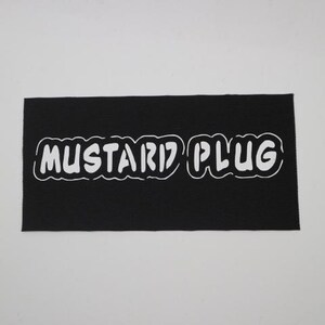 May include: A black rectangular patch with the white text "MUSTARD PLUG" in a stylized font. The patch has a textured surface and is set against a white background.