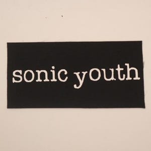 May include: A black rectangular patch with the words "sonic youth" in white, stylized lettering. The patch has a textured surface, and the text is centered horizontally. The background is a plain, off-white color.