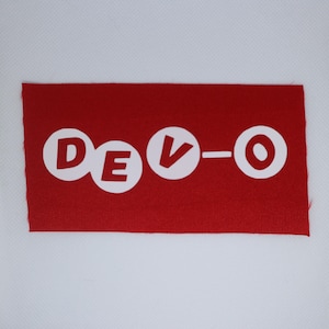 May include: A red rectangular patch featuring the white text "DEVO" in a circular font. The fabric has a textured appearance, and the edges are unfinished. The patch is set against a white background.