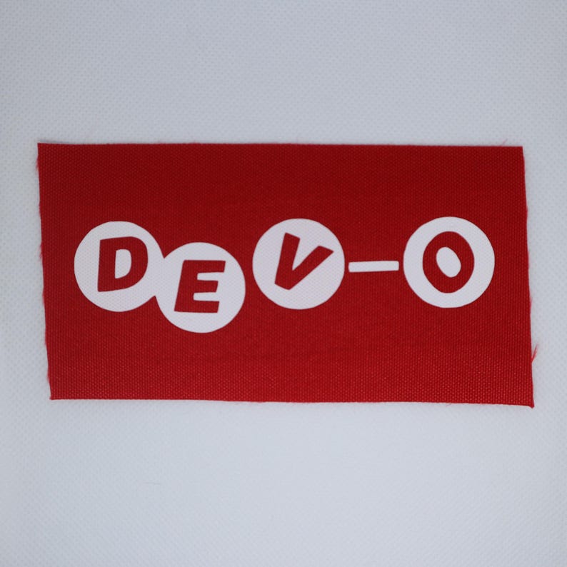 May include: A red rectangular patch featuring the white text "DEVO" in a circular font. The fabric has a textured appearance, and the edges are unfinished. The patch is set against a white background.