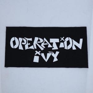 May include: A rectangular black fabric patch featuring the white text OPERATION IVY in a stylized, blocky font. The letters are arranged in two lines with the word OPERATION on the top and IVY on the bottom, centered on the black background.