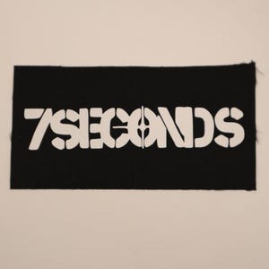 May include: A black rectangular patch with the words "7 SECONDS" in white, stencil-style lettering. The patch has a raw edge and is set against a neutral background. The design is simple and bold, suitable for apparel or accessories.