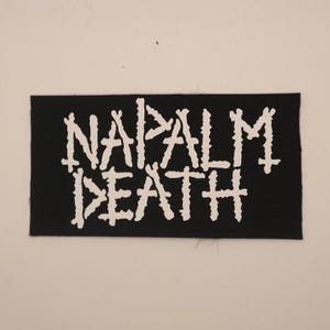 May include: A black rectangular patch with the words "NAPALM DEATH" in white, stylized, jagged lettering. The patch is likely for a jacket or bag, representing a band or a statement.