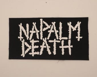 May include: A black rectangular patch with the words "NAPALM DEATH" in white, stylized, jagged lettering. The patch is likely made of fabric and is set against a neutral background.