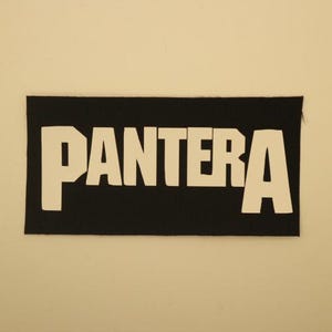 May include: A black rectangular patch with the word "PANTERA" in bold, white capital letters. The patch is likely made of fabric and is against a light beige background. The letters have a slightly distressed appearance.