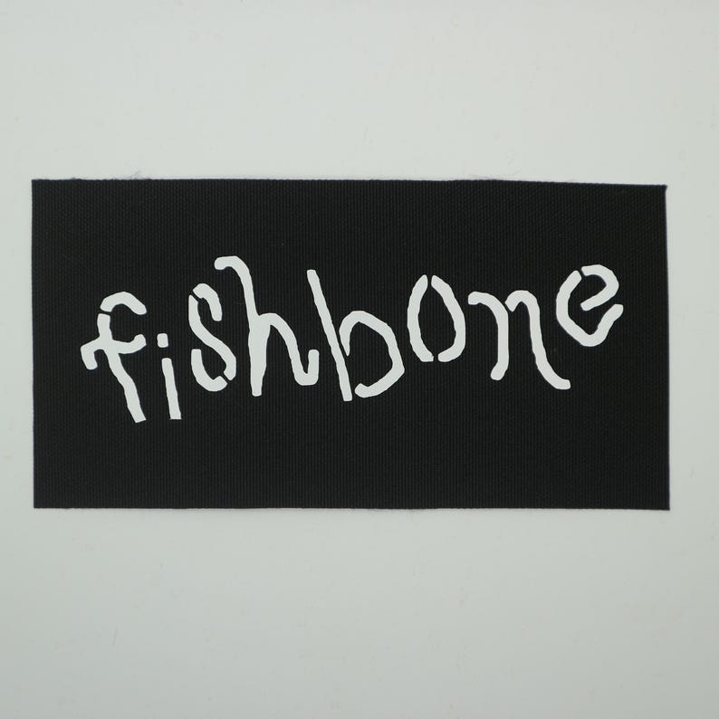 Op de afbeelding: Een zwart rechthoekig embleem met het woord "fishbone" in witte, sjabloonachtige letters. Het embleem heeft een getextureerd oppervlak en is waarschijnlijk gemaakt van stof.