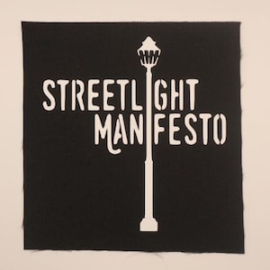 May include: Black square patch with the words "STREETLIGHT MANIFESTO" in white, along with a white illustration of a street lamp. The patch has a textured appearance and a raw edge.