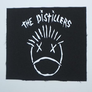 May include: Black square patch with white text and a graphic of a face. The text reads "THE DISTILLERS" above a stylized face with crossed-out eyes and a downturned mouth. The patch has a raw edge.