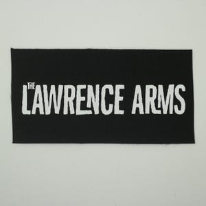 May include: A black rectangular patch with the white text "THE LAWRENCE ARMS". The text is in a bold, slightly distressed font. The patch has a ribbed texture and is set against a white background.