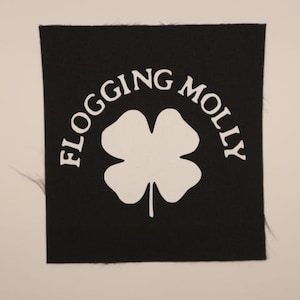 May include: A square black fabric patch featuring a white four-leaf clover graphic centered in the middle. The text FLOGGING MOLLY is printed in a white arched font above the clover. The edges of the patch show slight fraying of the fabric.
