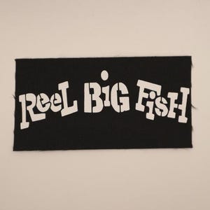 May include: Black rectangular patch with the words "Reel Big Fish" in white stencil-style lettering. The patch is made of a textured fabric and is set against a plain white background.