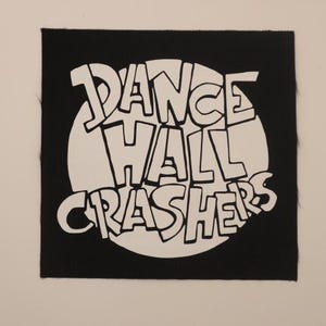 May include: Square black patch with the words "DANCE HALL CRASHERS" in white, stylized lettering. The text is arranged in a circular pattern, with the words inside a white circle. The patch is likely for clothing or accessories.