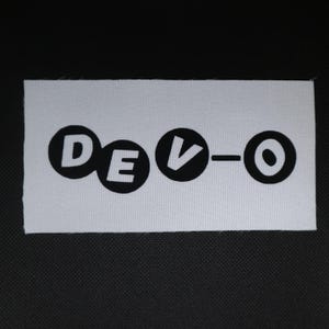 May include: A white rectangular patch with the word "DEVO" in black, stylized lettering. The letters are in a rounded font, with the "V" featuring a downward-pointing arrow. The patch is likely for clothing or accessories.