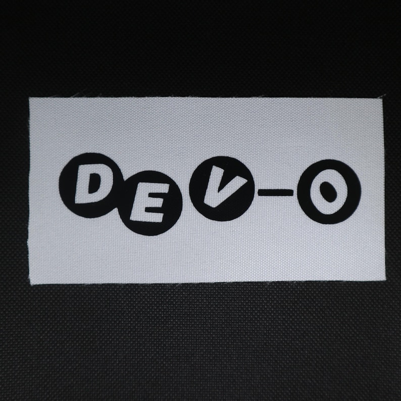 May include: A white rectangular patch featuring the word "DEVO" in black, rounded lettering. The "V" has a downward-pointing arrow. The patch is likely for clothing or accessories, and is made of a woven fabric.