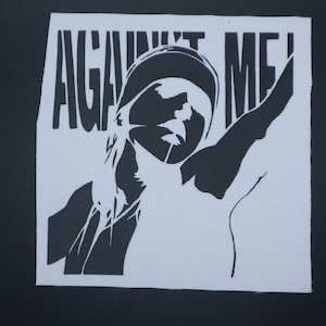 May include: A white square patch with the words "AGAINST ME!" in bold, black capital letters. A black and white illustration of a person with their arm raised is below the text. The patch is on a black background.