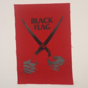 May include: A rectangular red fabric patch featuring a black graphic print. The design displays the text BLACK FLAG above two crossed black bars, with two stylized black hands positioned at the bottom, each gripping one of the crossed bars.