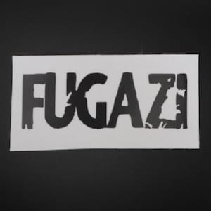 May include: A white rectangular patch with the word "FUGAZI" in bold, black, distressed lettering. The patch is set against a black background. The design is simple and graphic.