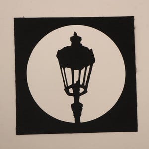 May include: A black and white patch featuring a silhouette of a vintage street lamp. The lamp is centered within a white circle, set against a black square background. The design is simple and graphic.
