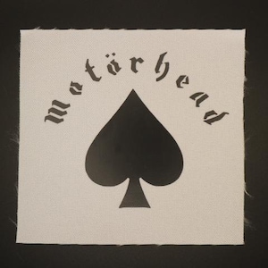 May include: Square, off-white patch featuring the word "Motörhead" arched above a black spade symbol. The text is in a gothic font. The patch has a textured surface and a raw edge.