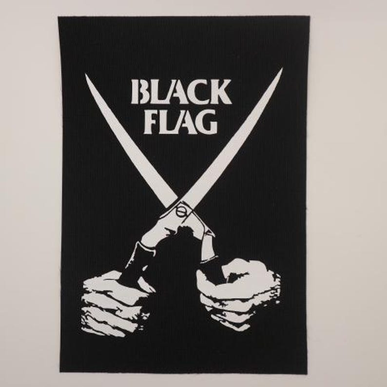 May include: A rectangular black fabric patch featuring a white screen-printed graphic of two hands holding a pair of open scissors. The text "BLACK FLAG" is printed in white capital letters above the scissors, centered on the black background.