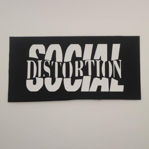 May include: A black rectangular patch with the words "SOCIAL DISTORTION" in white, stylized, uppercase letters. The text is arranged in three lines. The patch has a woven texture.