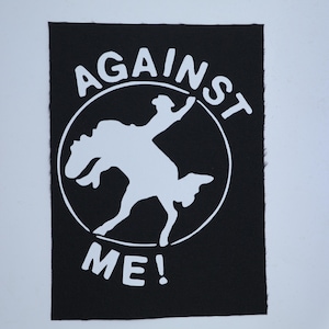 May include: Black patch with white text and graphic. The text reads "AGAINST ME!" in a circular design. The graphic is a silhouette of a person riding a bucking bronco.