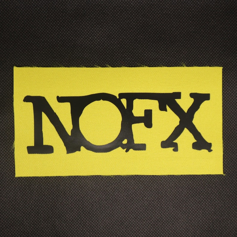 Peut inclure: Un &eacute;cusson rectangulaire jaune avec le texte noir "NOFX". Les lettres sont en gras et stylis&eacute;es. L'&eacute;cusson est fait d'un tissu textur&eacute;, sur un fond gris fonc&eacute;.