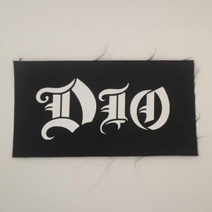 May include: Black rectangular patch with the word "DIO" in white gothic lettering. The patch has a textured surface and frayed edges, set against a plain white background. The design is simple and bold.
