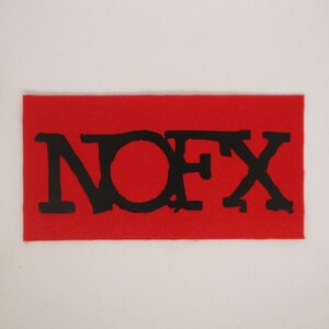 Peut inclure: Un &eacute;cusson rectangulaire rouge avec les lettres noires "NOFX" imprim&eacute;es dessus. Les lettres sont dans une police en gras, l&eacute;g&egrave;rement vieillie. L'&eacute;cusson a un aspect textur&eacute;.