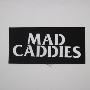 May include: A black rectangular patch with the white text "MAD CADDIES" in a bold, sans-serif font. The patch is likely made of fabric and is set against a plain white background. The text is centered on the patch.
