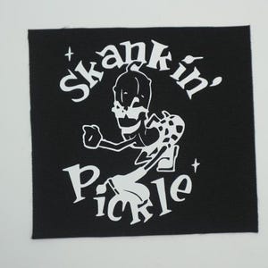 May include: Black square patch with white text and a cartoon skeleton figure. The text reads "skankin' pickle" in a stylized font. The skeleton is dancing with one arm raised. The patch has a textured appearance.