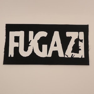 May include: A black rectangular patch with the word "FUGAZI" in large, distressed white letters. The patch has a textured appearance and a simple, bold design, suitable for clothing or accessories.
