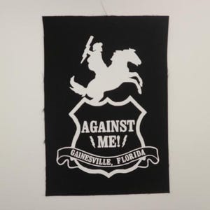 May include: Black and white embroidered patch with a shield design. The shield features a silhouette of a woman riding a horse, with the text "AGAINST ME!" and "GAINESVILLE, FLORIDA" below.