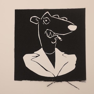 May include: A black square patch featuring a white cartoon illustration of a dog-like character wearing a blazer and smoking a cigarette. The character has a wide grin and is outlined in white.