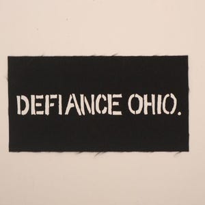 May include: A black rectangular patch with the words "DEFIANCE OHIO." in white, stencil-style lettering. The patch has a slightly frayed edge and is set against a neutral background.