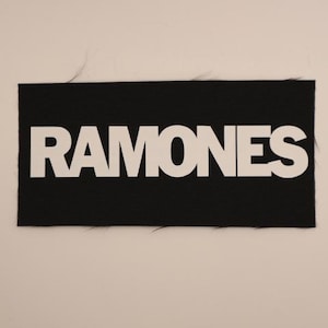 May include: Black rectangular patch with the word "RAMONES" in bold white capital letters. The patch has a textured surface and frayed edges, set against a neutral background. The text is centered on the patch.