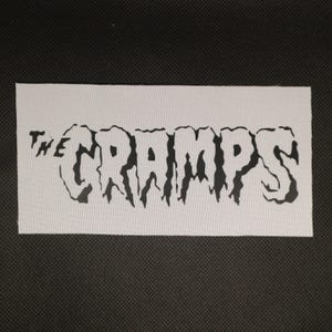 May include: A rectangular white patch with the words "THE CRAMPS" in a black, stylized font. The letters have a dripping, jagged appearance. The patch is set against a dark, textured background.