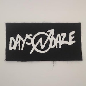 May include: Black rectangular patch with the white text "DAYS @ DAZE" in a stylized font. The text is centered on the patch. The patch has a textured surface.