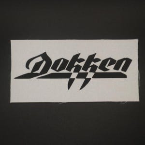 May include: A rectangular white patch with the black word "Dokken" in a stylized font. The patch has a textured surface and is set against a black background. The design includes sharp, angular elements.