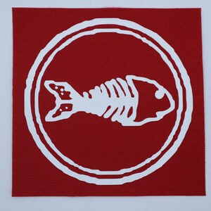 Puede incluir: Un parche cuadrado rojo con un dise&ntilde;o de esqueleto de pez blanco dentro de un borde circular blanco. El esqueleto del pez es una ilustraci&oacute;n detallada y el parche tiene una apariencia texturizada.