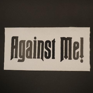 May include: A rectangular patch with the text "Against Me!" in a gothic font. The text is in shades of gray and black against a white background. The patch has a raw edge and is set against a black background.