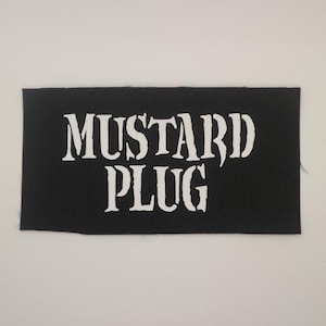 May include: Black rectangular patch with the words "MUSTARD PLUG" in large, white, stylized lettering. The patch is made of a textured fabric.