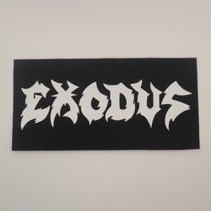May include: Black rectangular patch with the word "EXODUS" in white, stylized, jagged lettering. The patch has a textured surface and is likely made of fabric. The design is bold and eye-catching.
