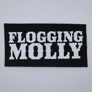 May include: A black rectangular patch with the words "FLOGGING MOLLY" in bold, white, block letters. The patch is likely for a band or merchandise.