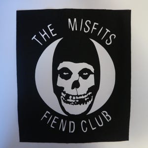 May include: Black square patch featuring a white skull design within a white crescent moon shape. The text "THE MISFITS" arches above the skull, and "FIEND CLUB" is below. The patch is likely for clothing or accessories.